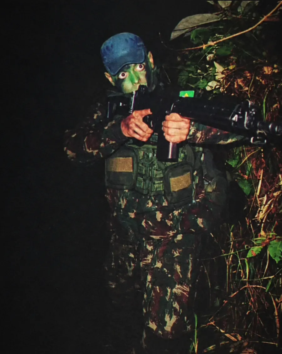 Guilherme Carvalho de Andrade em operação noturna do Exército Brasileiro, com camuflagem no rosto, colete tático e fuzil, entre a vegetação.